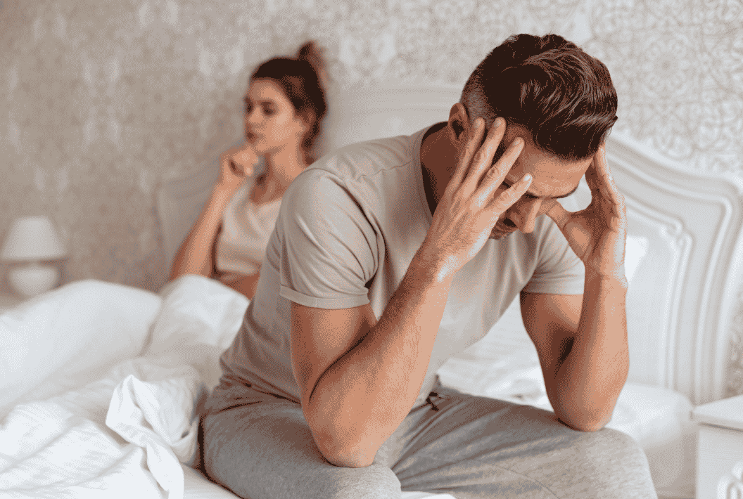 Distressed man sitting on the edge of a bed with his head in his hands while his partner lies behind him, representing anxiety-related sexual performance concerns.