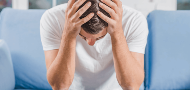 Man sitting on a sofa with his head in his hands, representing the emotional impact of Peyronie's disease.
