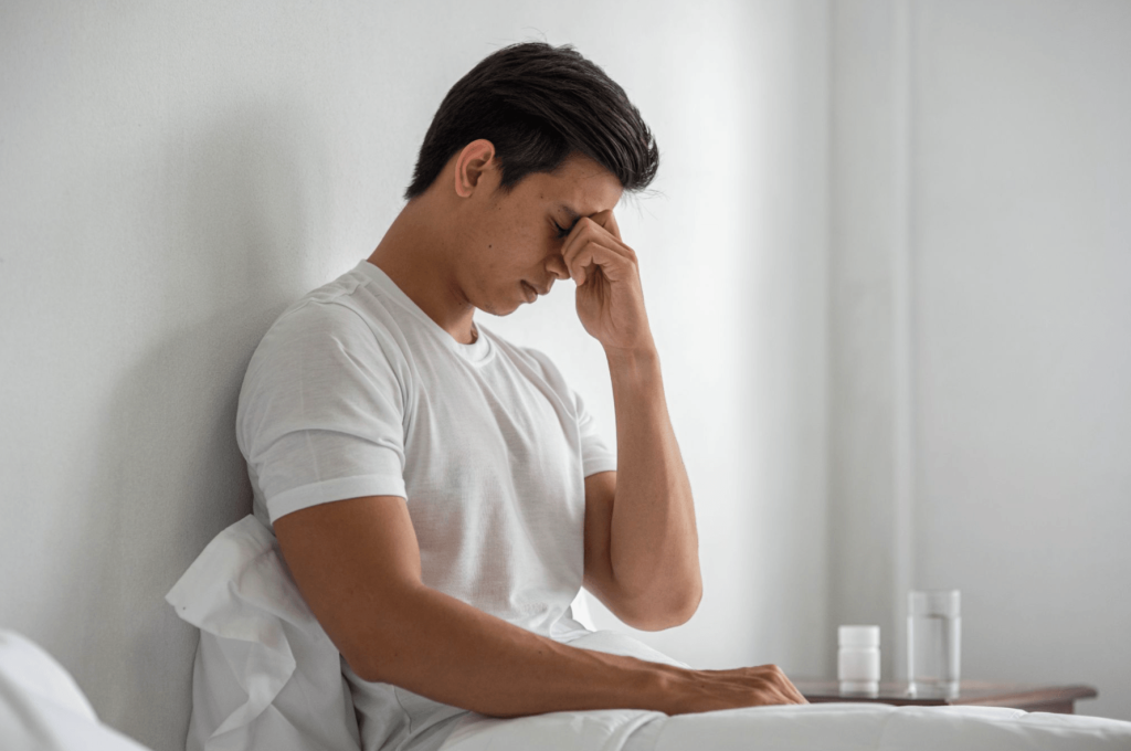 Man sitting on a bed with his head in his hand, illustrating tiredness and symptoms linked to low testosterone or hormonal changes.